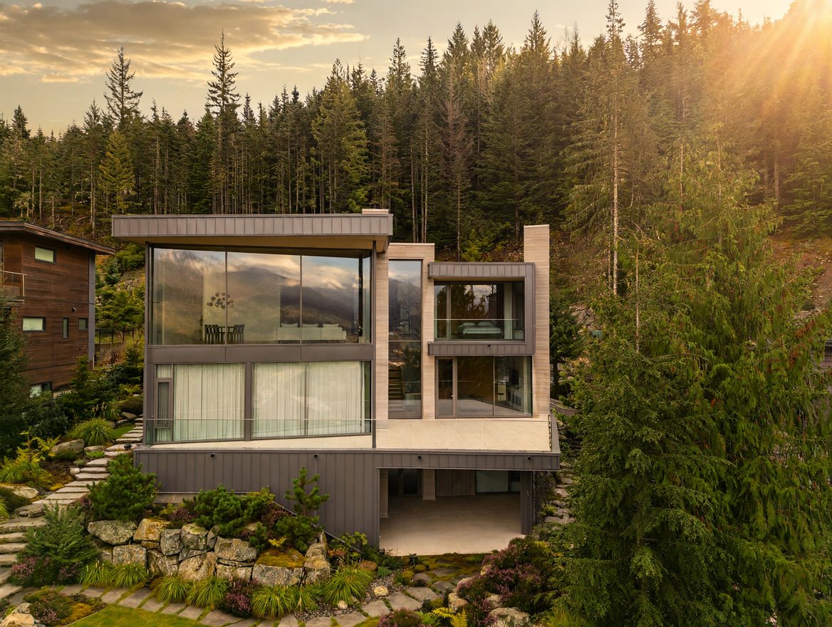 Contemporary glass-walled residence in warm golden-hour light with completed stonework and layered evergreen landscaping